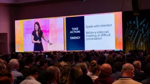 Allison Shapira speaking on stage to a large audience at a leadership conference, with a presentation slide behind her that reads “TAKE ACTION: ENERGY – Speak with Intention.
