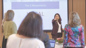 Allison Shapira leading a workshop, pointing toward participants standing in front of a presentation slide titled "The 3 Movements."