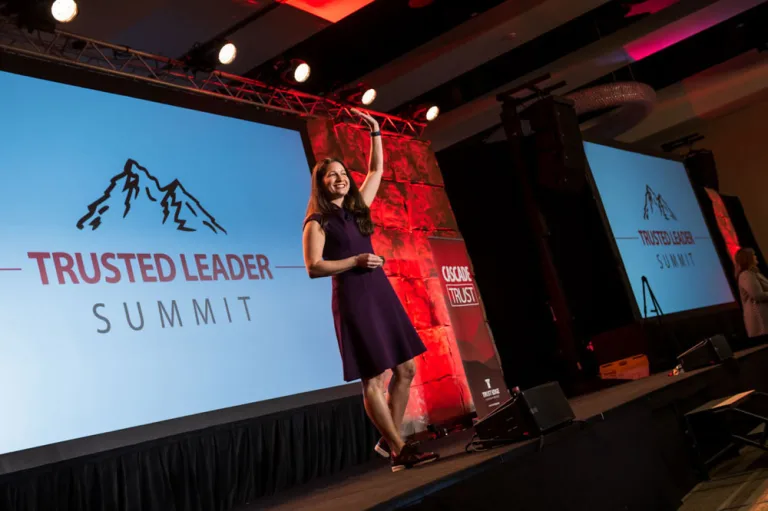Allison Shapira speaking on stage at the Trusted Leader Summit, smiling and gesturing to the audience.