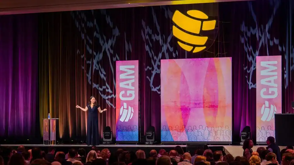 Allison Shapira speaks to a large audience from the stage at a corporate conference.