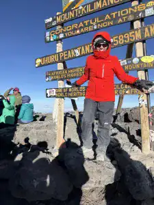 Allison Shapira stands at Uhuru Peak, the summit of Mount Kilimanjaro in Tanzania, wearing a bright red jacket in front of the summit sign.